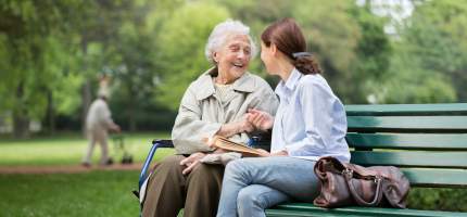 Eine Seniorin sitzt mit Ihrer Pflegerin auf einer Parkbank.