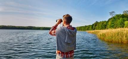 Ein junger Mann schaut mit einem Fernglas auf einen See.