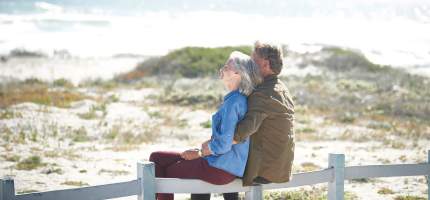 Ein älteres Ehepaar sitzt gemeinsam auf einem Zaun in den Dünen am Strand.