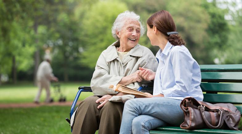 Eine Seniorin sitzt mit Ihrer Pflegerin auf einer Parkbank.